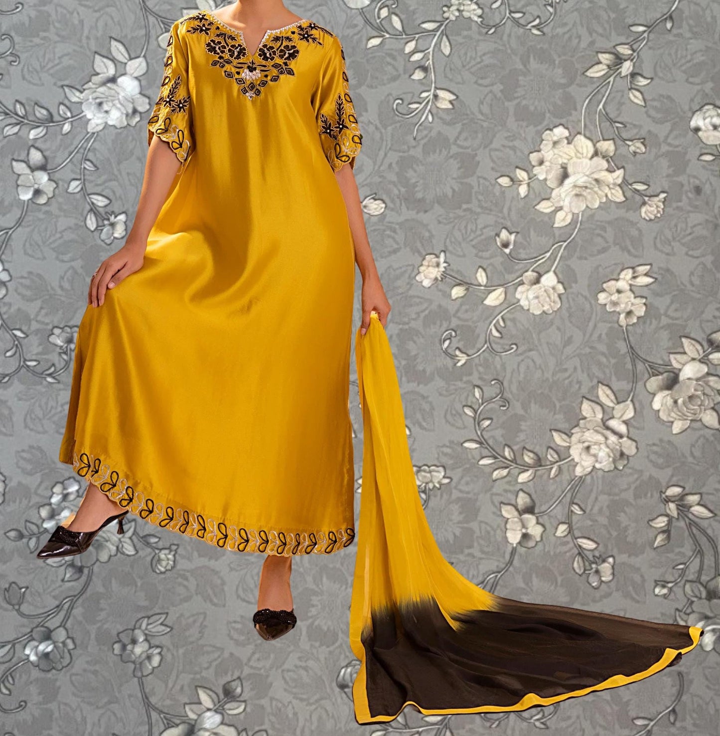 A woman wearing a long, yellow dress with gold embroidery and black heels is standing against a gray floral background.