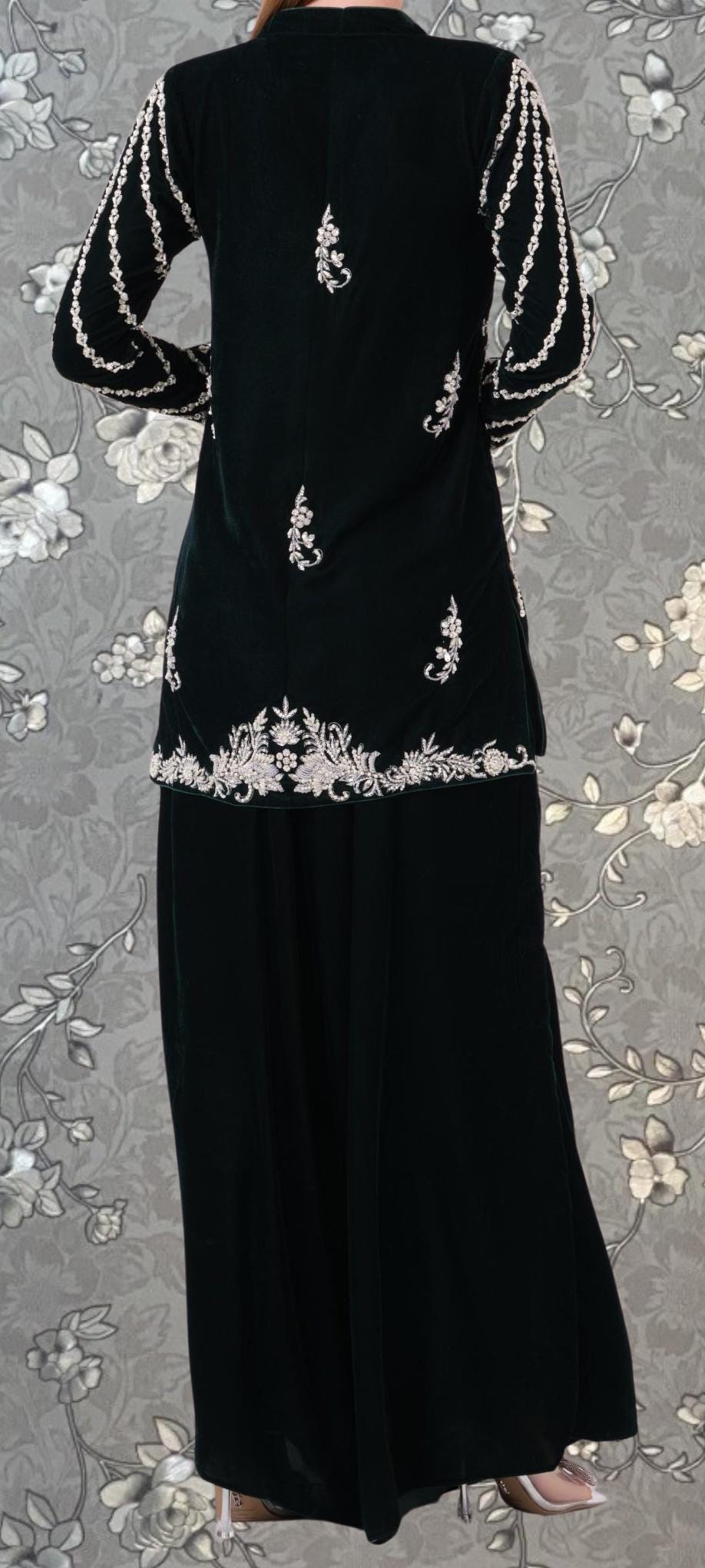 A person wearing a black dress with white embroidered designs on the sleeves and skirt, standing against a floral patterned background.