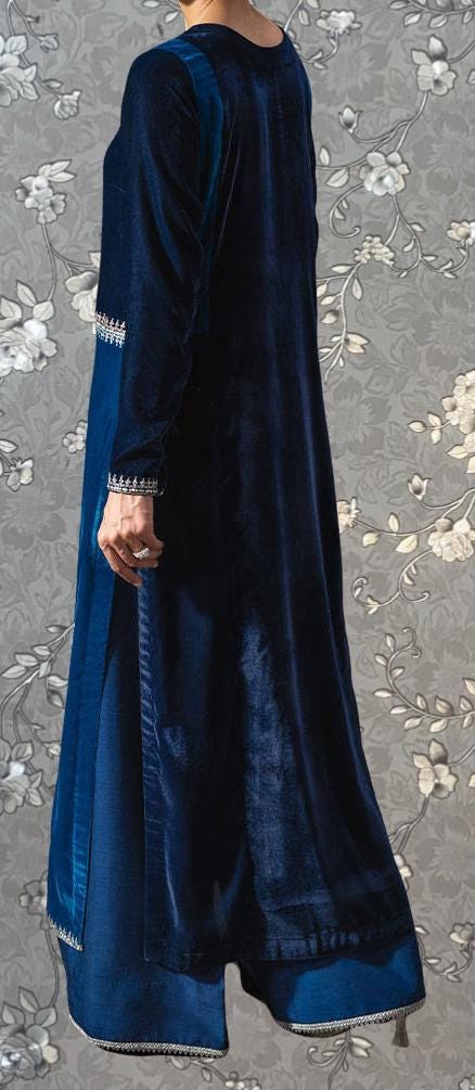 A person wearing a long, blue velvet dress with a high collar and long sleeves stands against a gray floral background.