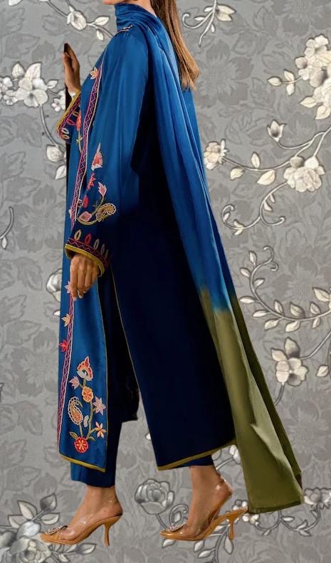 A person wearing a long blue and green dress with intricate embroidery, standing against a gray floral background.