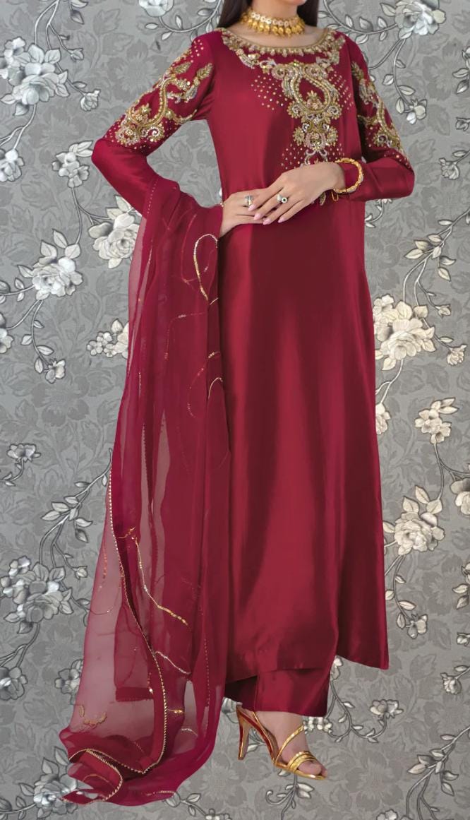 A woman wearing a long, burgundy dress with gold embroidery and a matching red shawl. She is posing with her hands on her hips, and the background features a gray floral pattern.