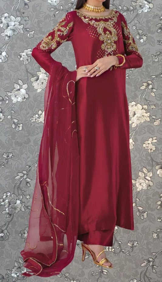 A woman wearing a long, burgundy dress with gold embroidery and a matching red shawl. She is posing with her hands on her hips, and the background features a gray floral pattern.