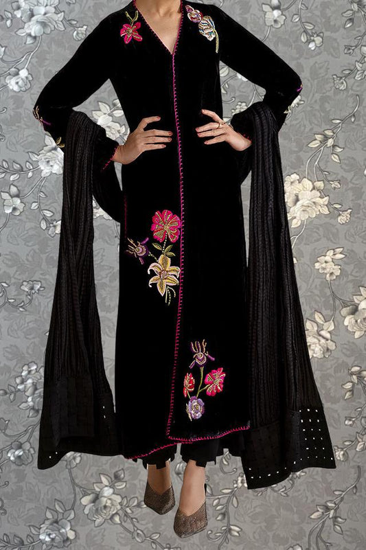 A person wearing a long black dress with pink and yellow floral embroidery, standing against a gray floral background.