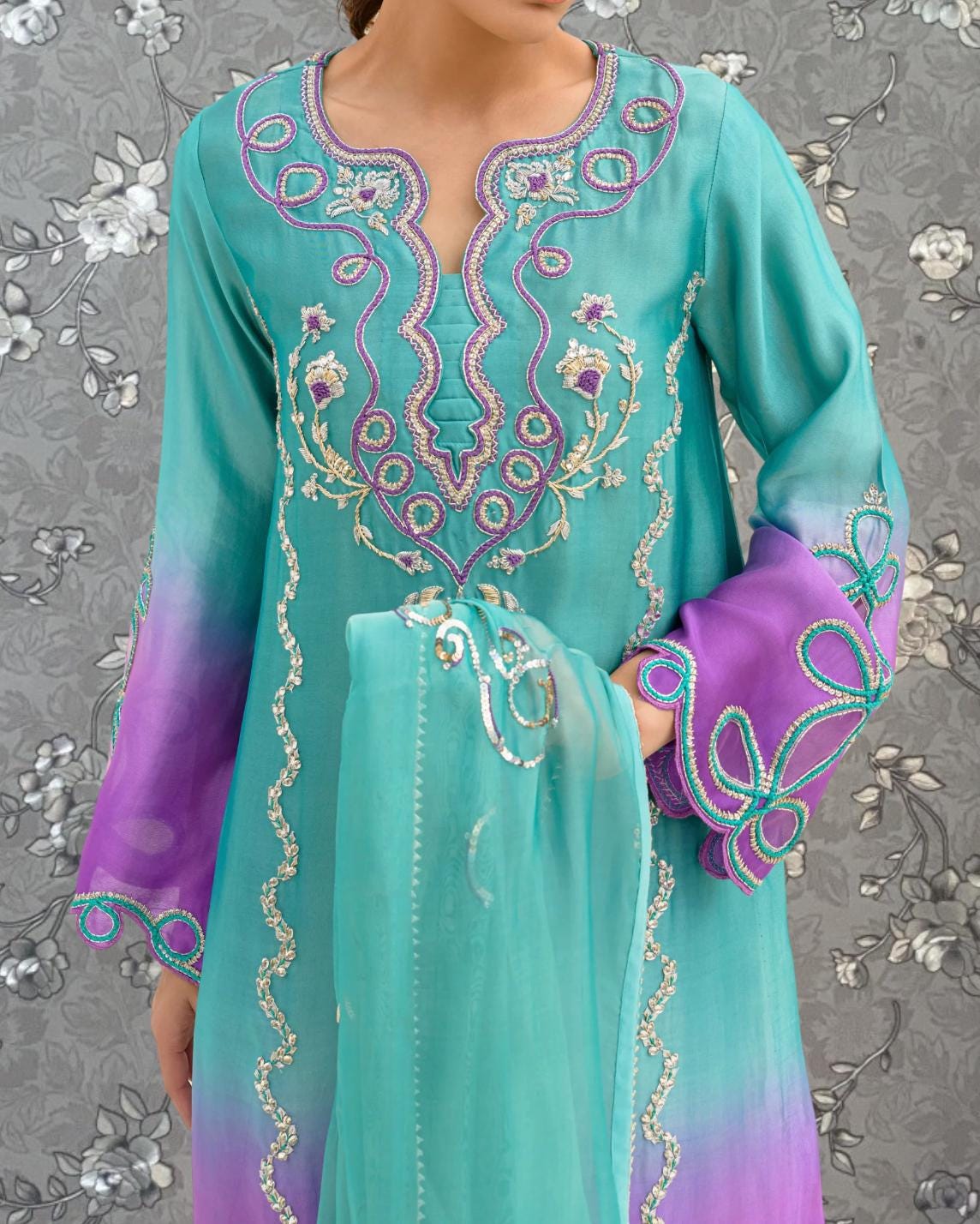 A woman wearing a turquoise and purple dress with intricate embroidered patterns on the sleeves and collar.