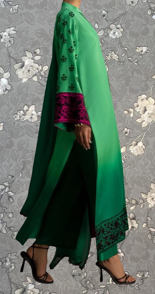 A person wearing a long green dress with pink accents and floral patterns, standing against a gray floral background.