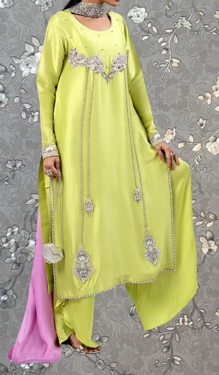 A woman wearing a bright yellow dress with intricate silver embroidery and dangling chains, posing against a gray floral background.