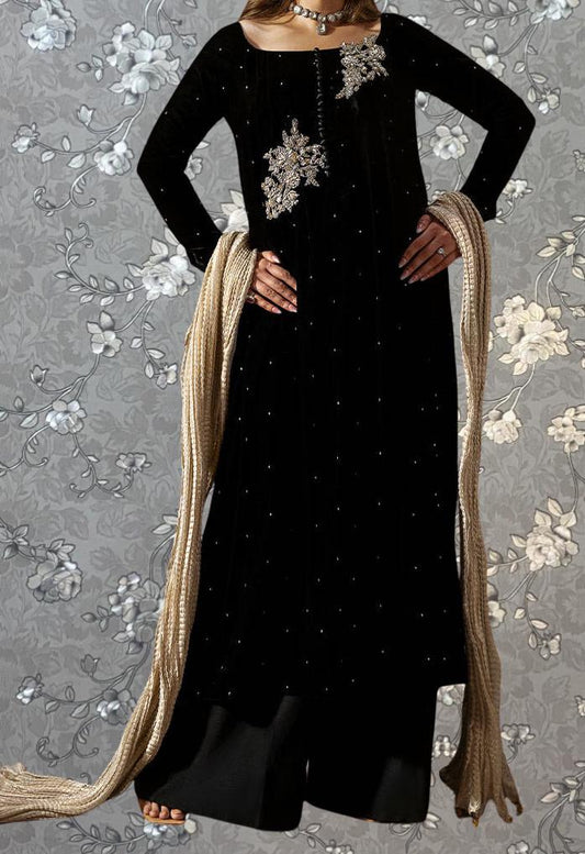 A woman wearing a black dress with gold embellishments and a long beige scarf, standing against a gray floral background.