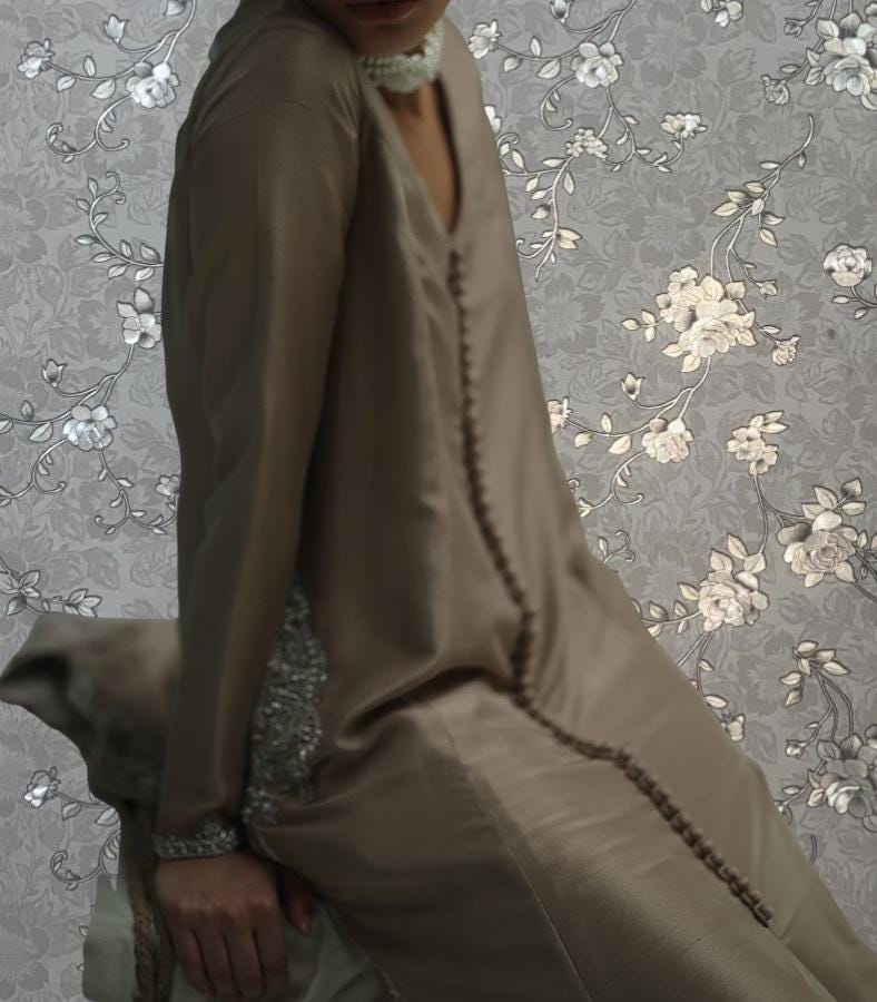 A woman wearing a long, beige dress with a floral pattern, posing against a gray floral background.