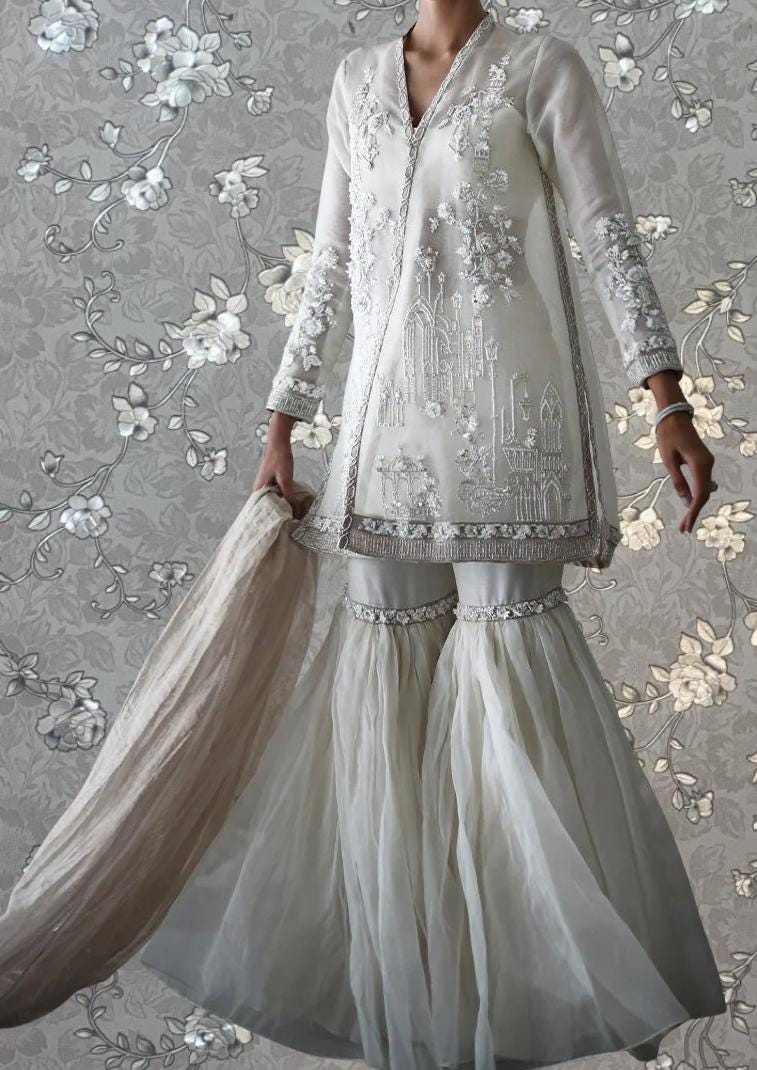 A woman wearing a white, long-sleeved dress with intricate embroidery and a high skirt, posing against a floral patterned background.