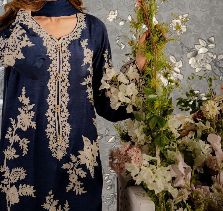 A woman wearing a blue dress with white floral embroidery stands next to a large floral arrangement.