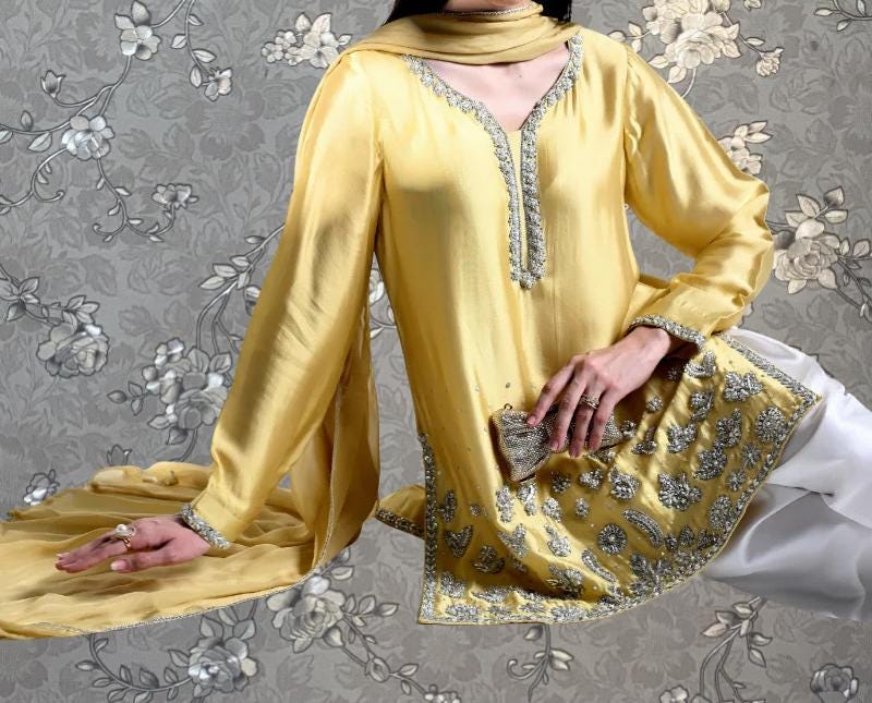 A woman wearing a yellow and white dress with intricate embroidery poses against a gray floral background.