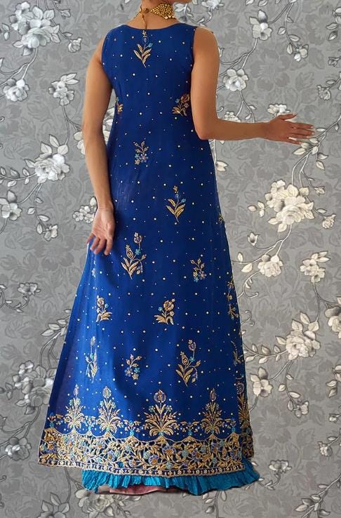 A woman wearing a long, blue dress with gold embroidery stands against a gray floral background.