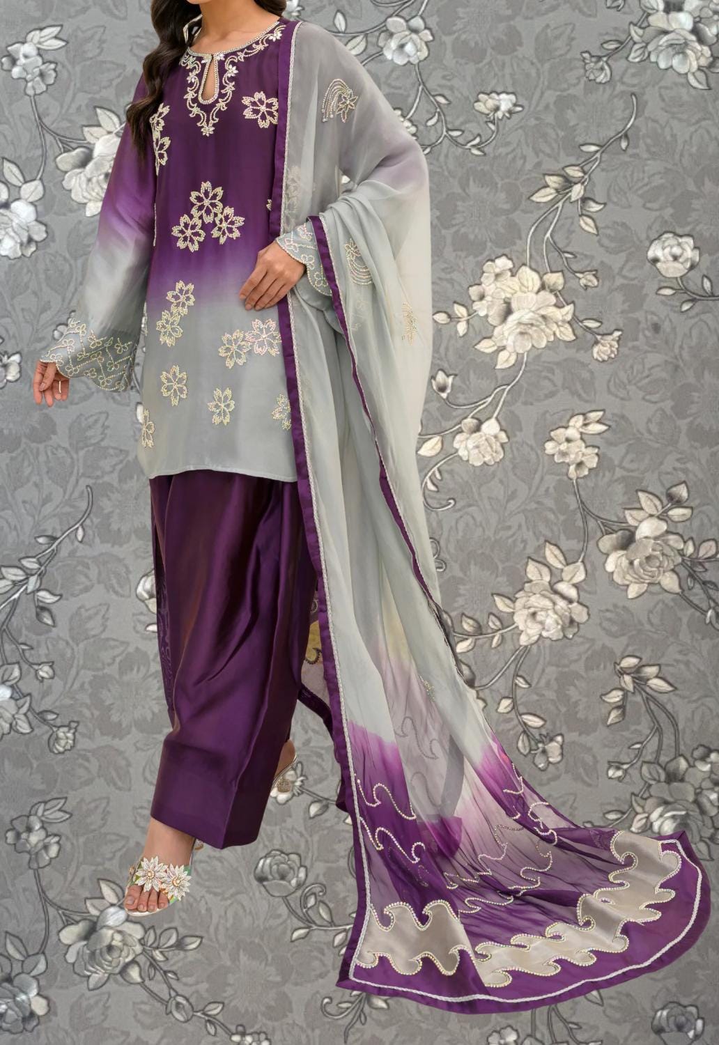 A woman wearing a purple and gray dress with a long purple shawl, standing against a gray floral background.