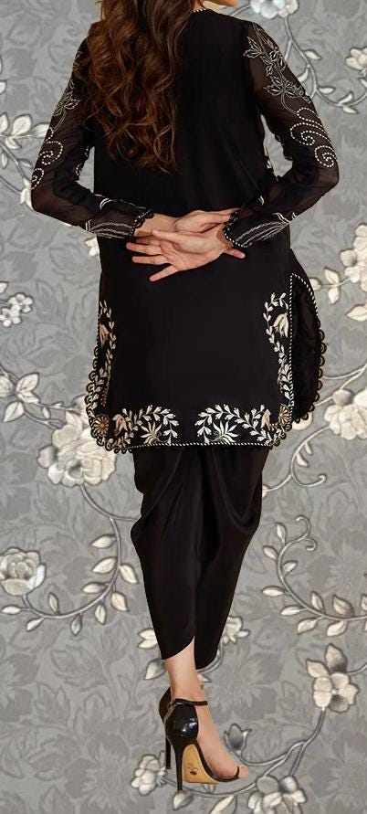 A woman wearing a black dress with intricate embroidery stands against a gray floral background.
