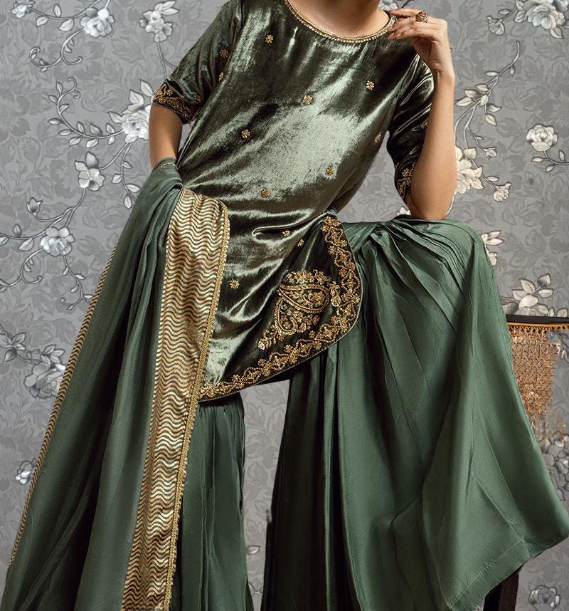 A person wearing a green velvet dress with gold embroidery and a matching green skirt, posing against a gray floral background.