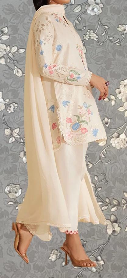 A woman wearing a long, white dress with floral embroidery stands against a gray floral background.