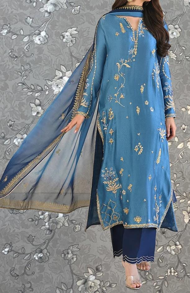 A woman wearing a blue and gold dress with intricate embroidery, standing against a gray floral background.