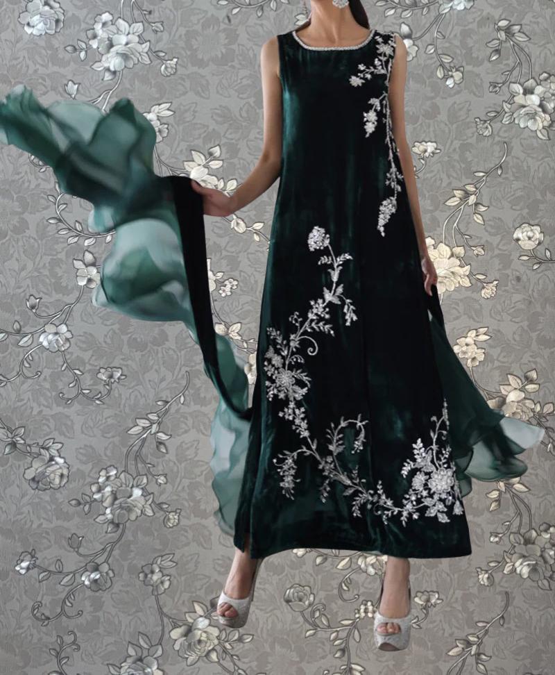 A woman wearing a long, green dress with white floral embroidery stands against a gray floral background.