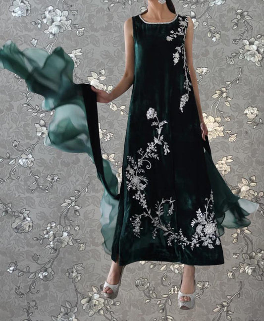 A woman wearing a long, green dress with white floral embroidery stands against a gray floral background.