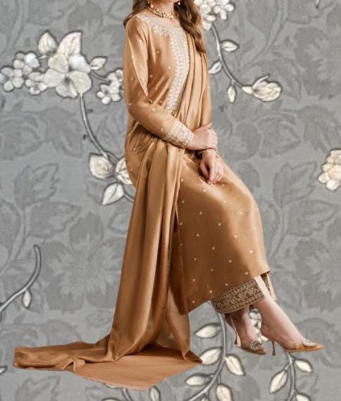 A woman wearing a long, gold-colored dress with lace trim and floral patterns, sitting on a gray background with white flowers.