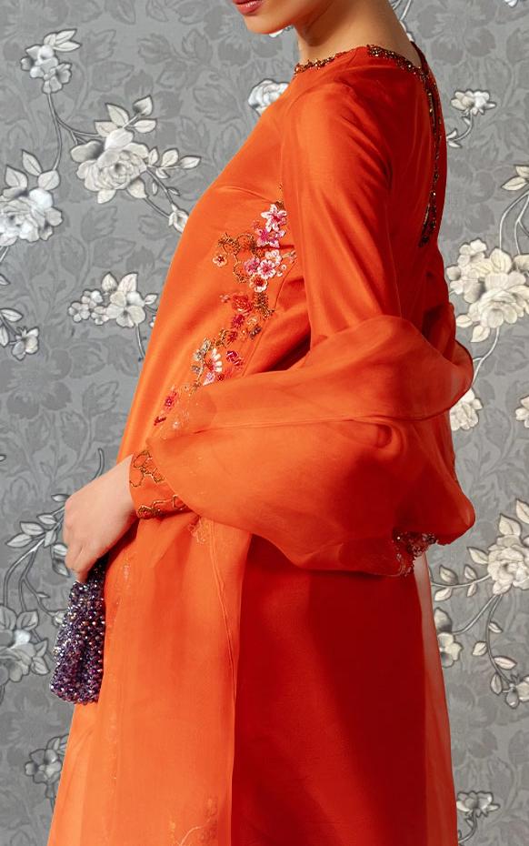 A woman wearing an orange dress with floral embroidery stands against a gray floral background.