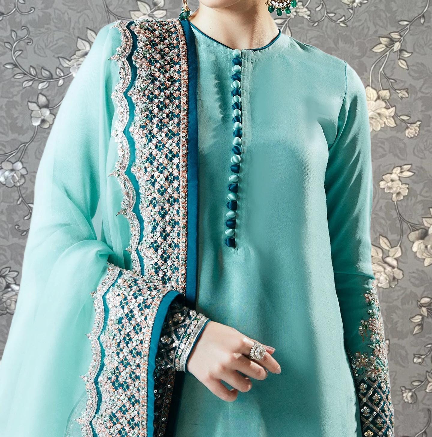 A woman wearing a turquoise dress with a matching turquoise shawl and a decorative blue and white patterned skirt. She is holding a ring on her finger.