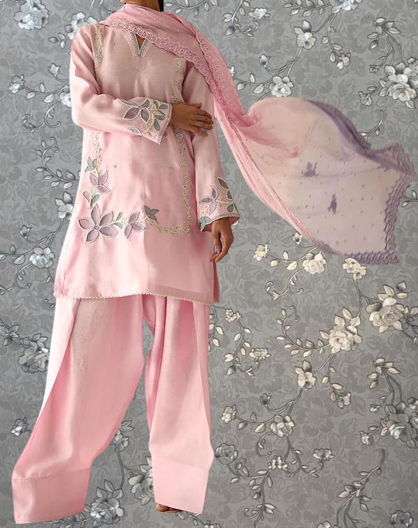 A person wearing a pink dress with floral embroidery and a matching pink shawl is shown against a gray floral background.
