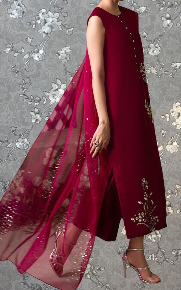 A person wearing a long, sleeveless red dress with a sheer, flowing skirt and high heels, posing against a gray floral background.