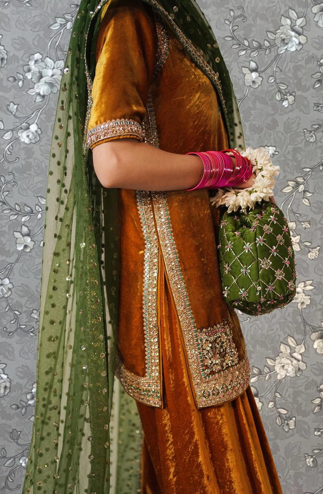 A person wearing a gold and green traditional Indian outfit, including a gold and green shawl, and holding a green and gold handbag with white flowers.