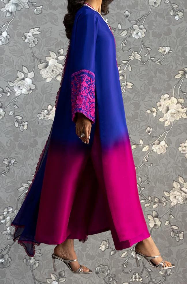 A woman wearing a long, flowing dress with a mix of blue and pink colors, standing against a gray floral background.