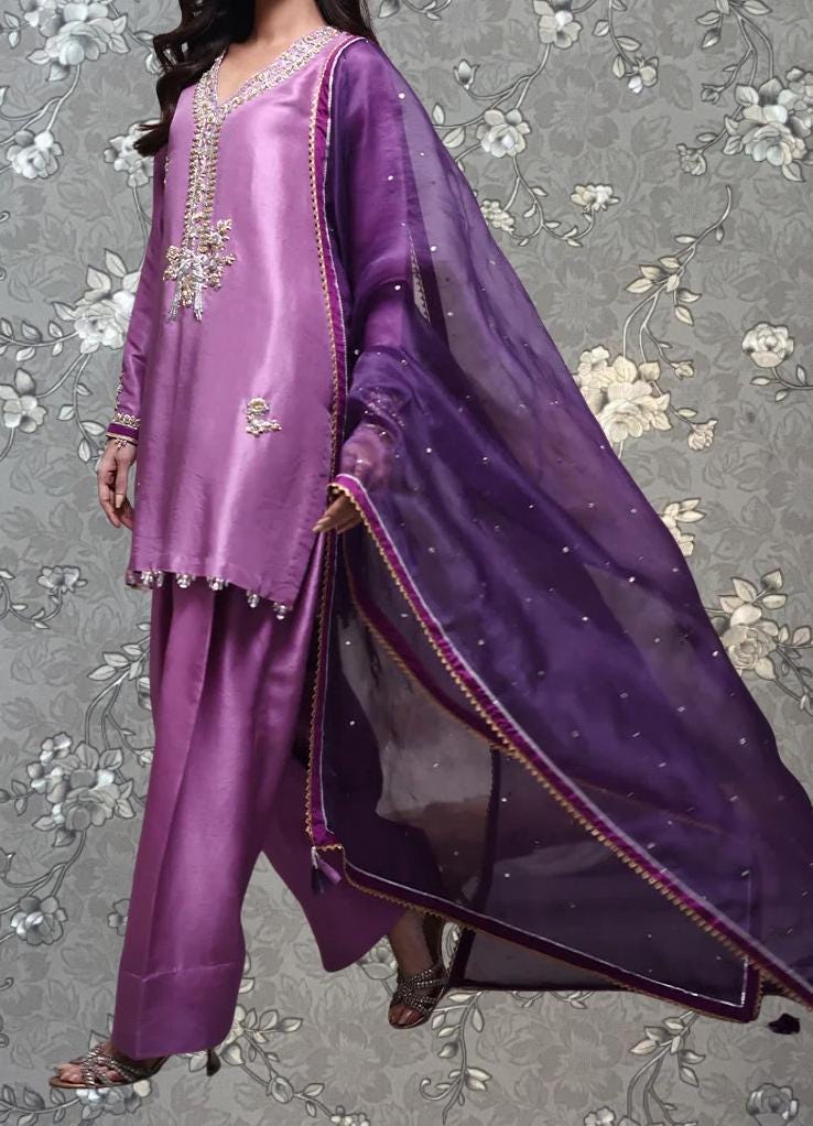 A woman wearing a purple dress with a matching purple shawl, standing against a gray floral background.