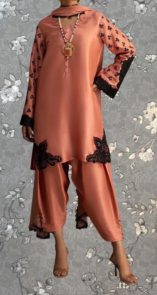 A woman wearing a peach-colored dress with black lace accents and floral patterns, standing against a gray floral background.