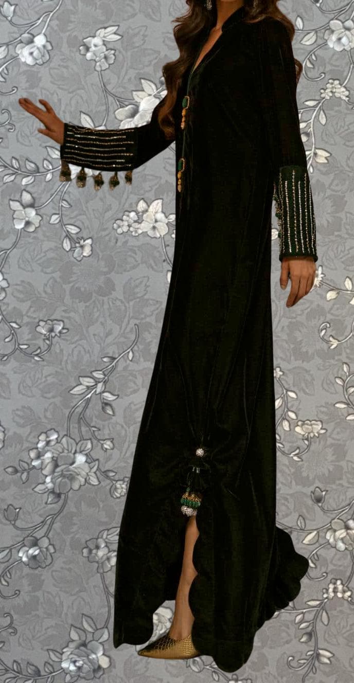 A person wearing a long black dress with gold embellishments and a high slit, standing against a gray floral background.