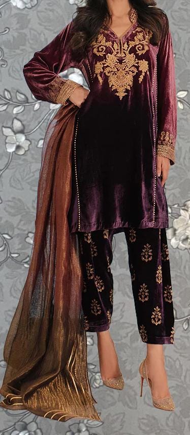 A woman wearing a purple velvet dress with gold embroidery and gold heels, standing against a gray floral background.