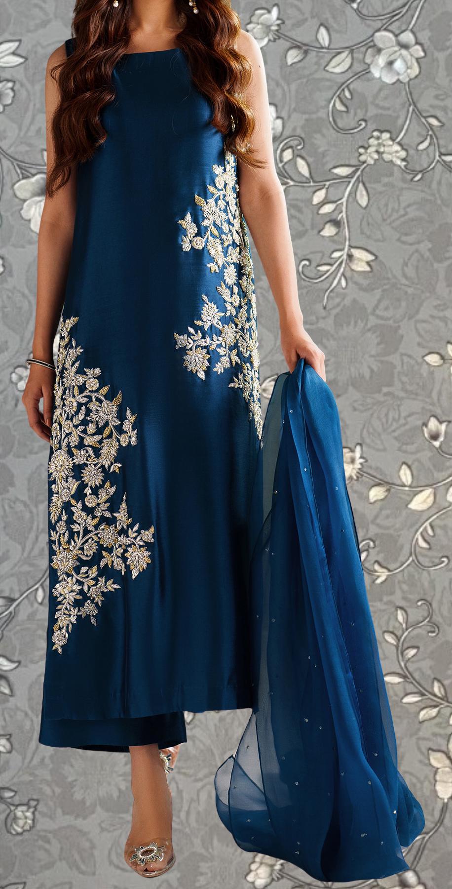 A woman wearing a blue dress with intricate embroidered designs, standing against a gray floral background.