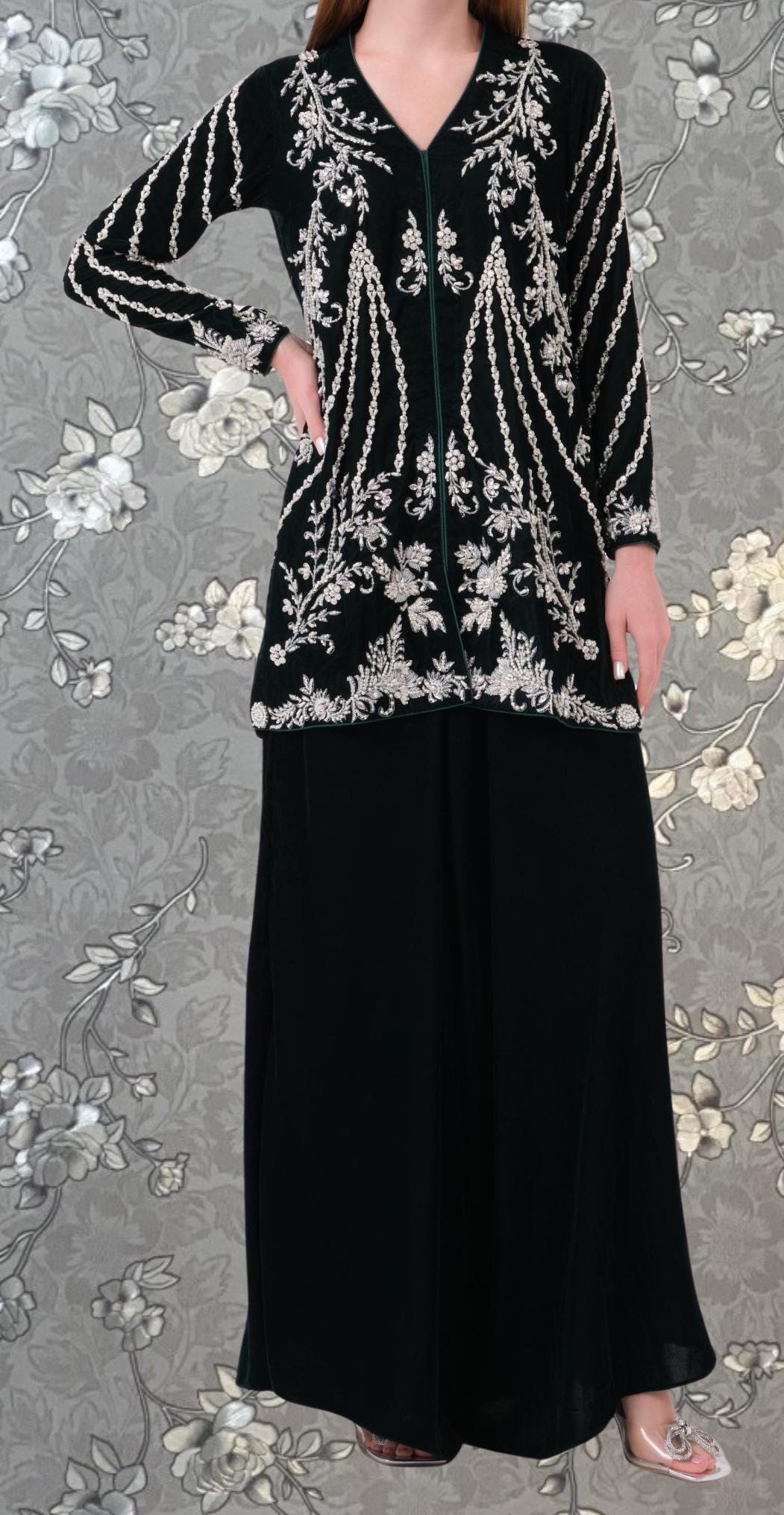 A woman wearing a black dress with white floral embroidery and a black jacket with white floral embroidery stands against a gray floral background.