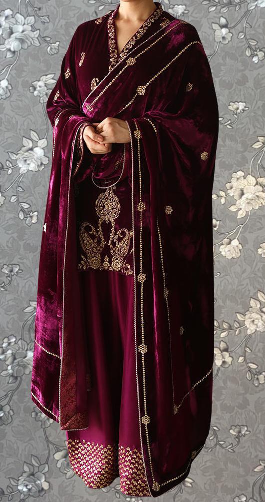A person wearing a long, burgundy velvet shawl with gold embroidery and a matching skirt, standing against a gray floral background.