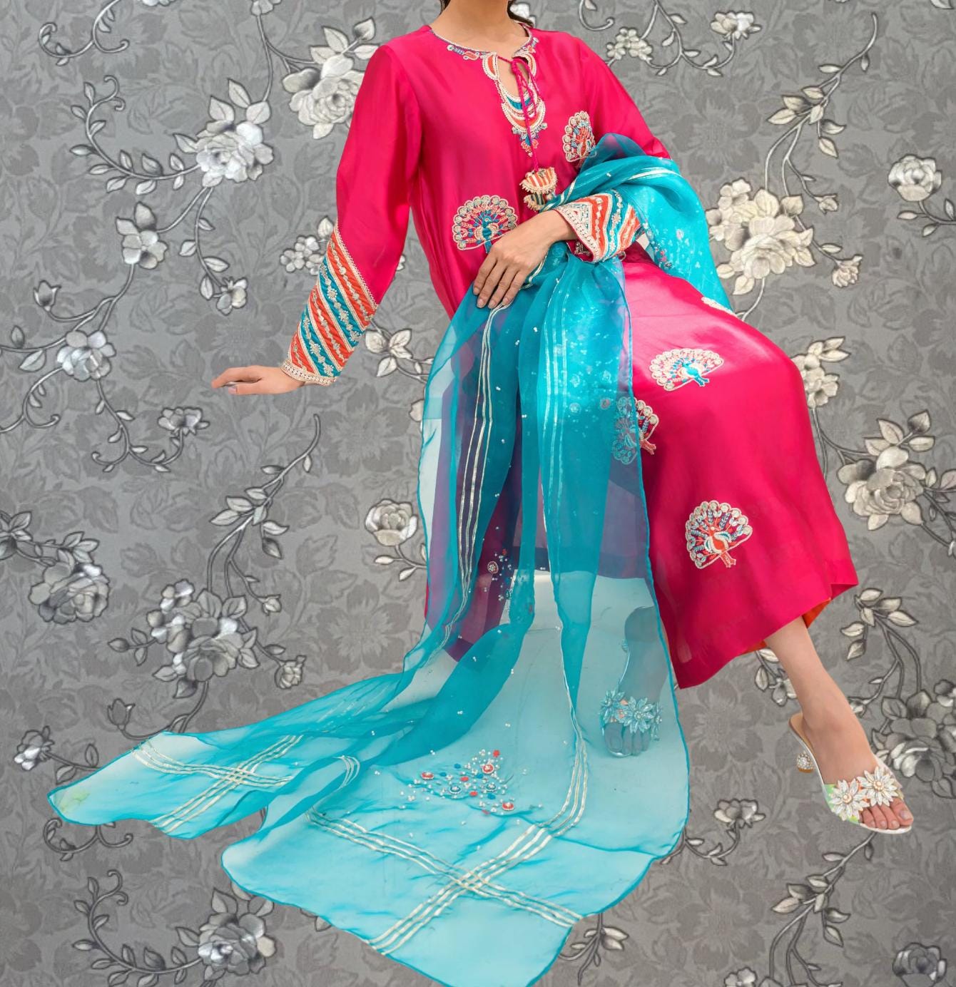 A woman wearing a vibrant pink and blue dress with intricate embroidery and a matching turquoise shawl, posing against a gray floral background.