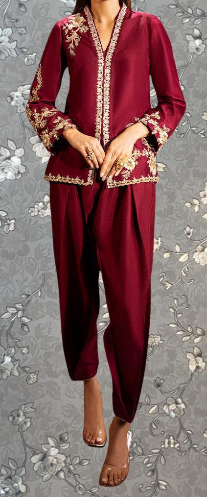 A woman wearing a red and gold outfit with intricate embroidery, standing against a gray floral background.