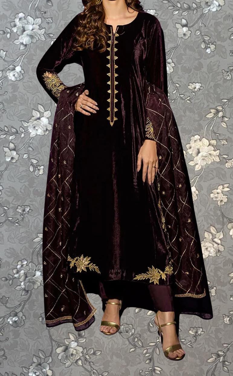 A woman wearing a long, burgundy velvet dress with gold embroidery and a matching shawl. She is posing against a gray floral background.