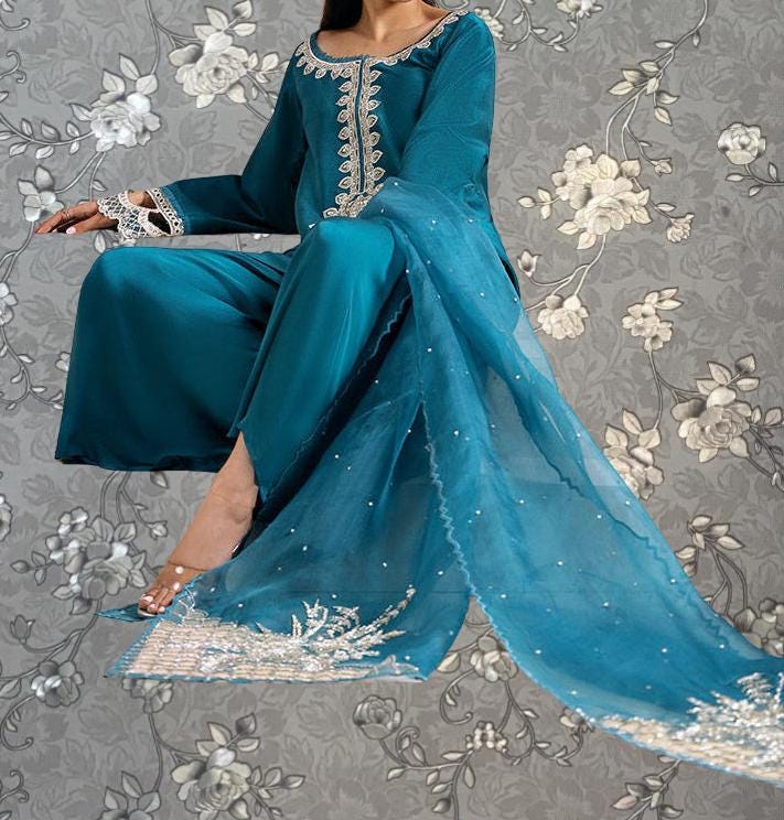 A woman wearing a teal blue dress with intricate embroidery and a matching blue shawl, sitting against a gray floral background.