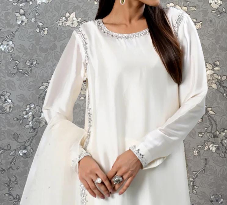 A woman wearing a white dress with a floral pattern and silver embellishments stands against a gray floral background.