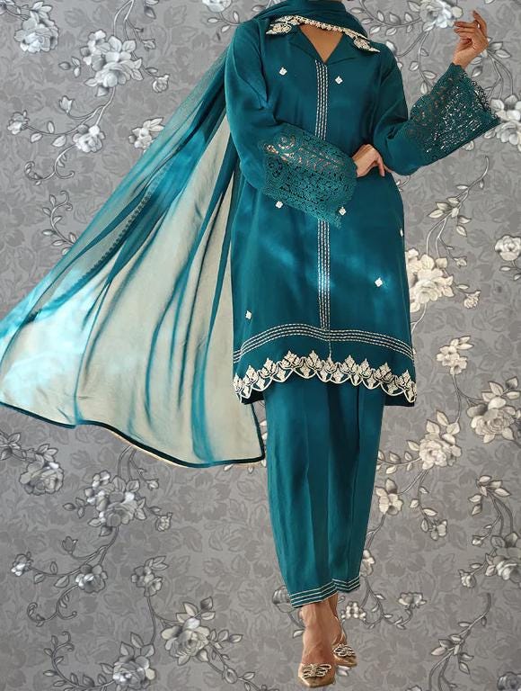 A person wearing a teal-colored dress with lace sleeves and a matching skirt, standing against a gray floral background.