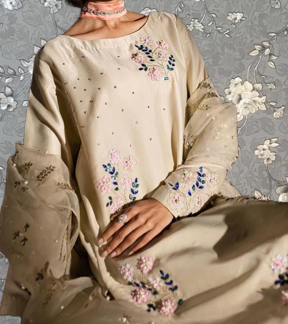 A woman wearing a beige dress with floral embroidery and a choker necklace is shown against a gray floral background.