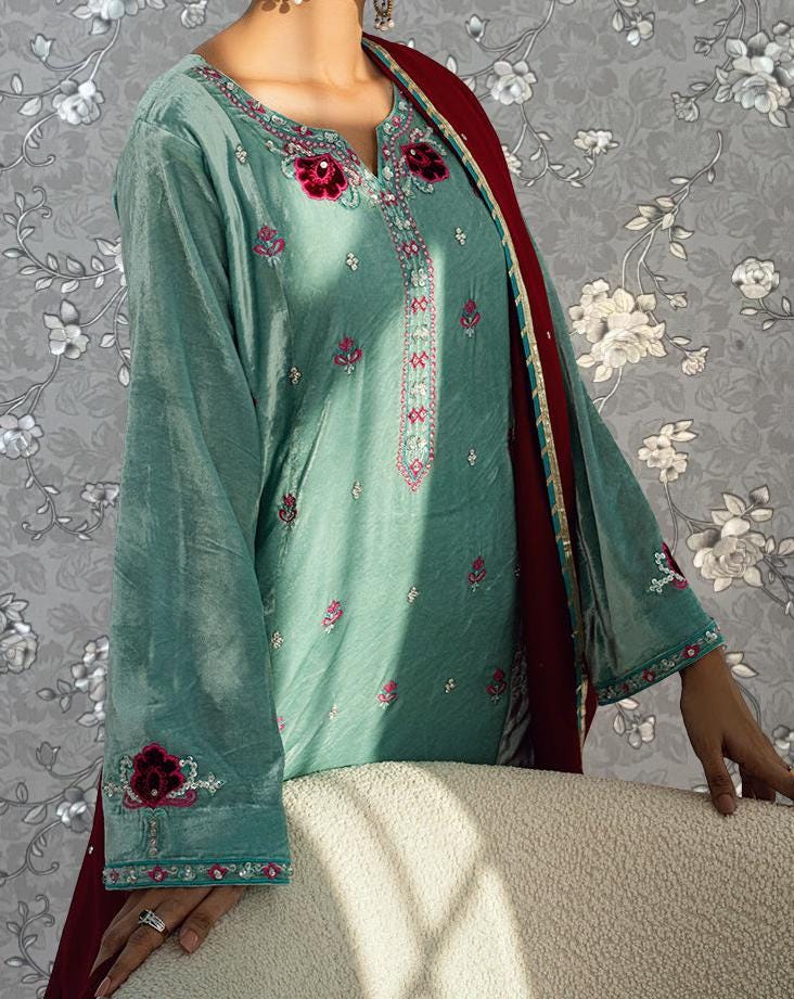 A woman wearing a green dress with red and pink floral embroidery stands against a gray floral background.