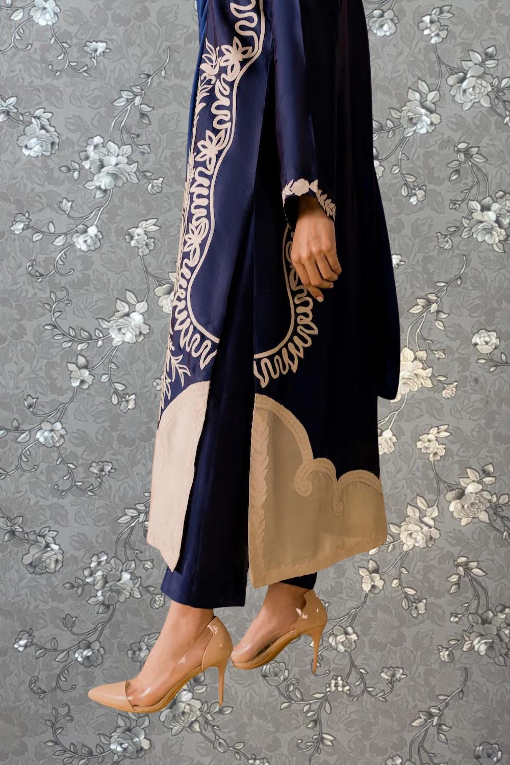 A person wearing a navy blue robe with gold embroidery and beige high heels stands against a gray floral background.
