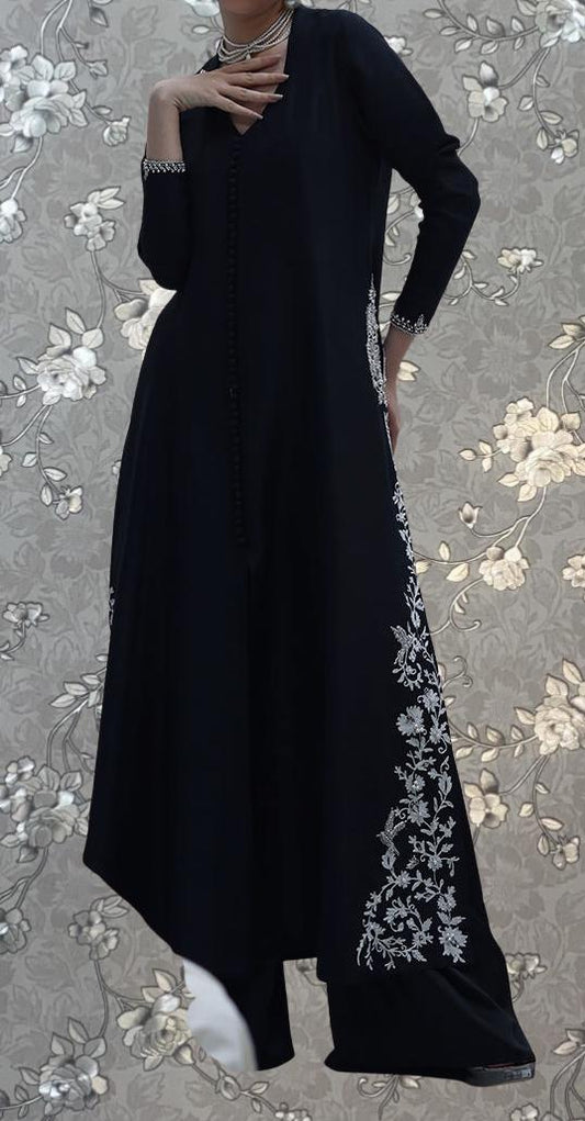 A person wearing a long black dress with white floral embroidery stands against a floral background.