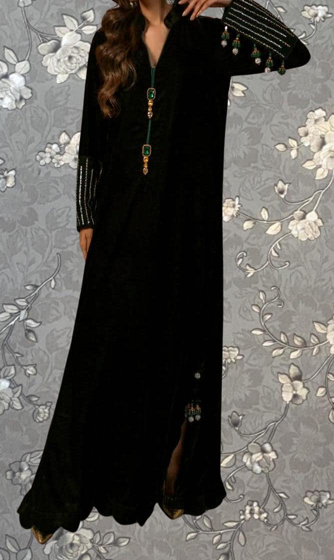 A woman wearing a long black dress with gold accents and green embellishments, posing against a gray floral background.