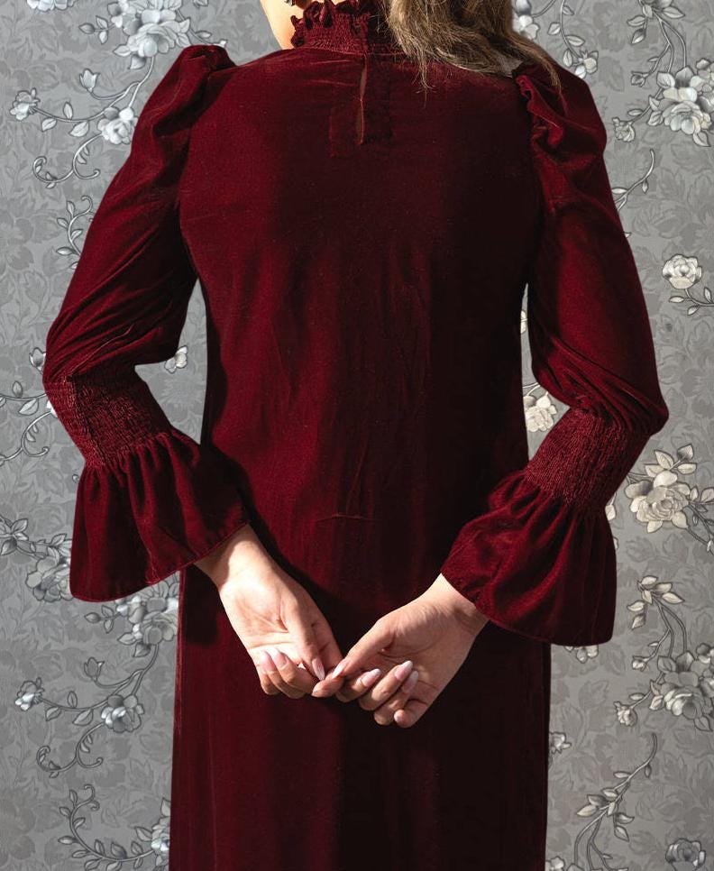 A person wearing a long, burgundy dress with puff sleeves stands in front of a floral patterned background.