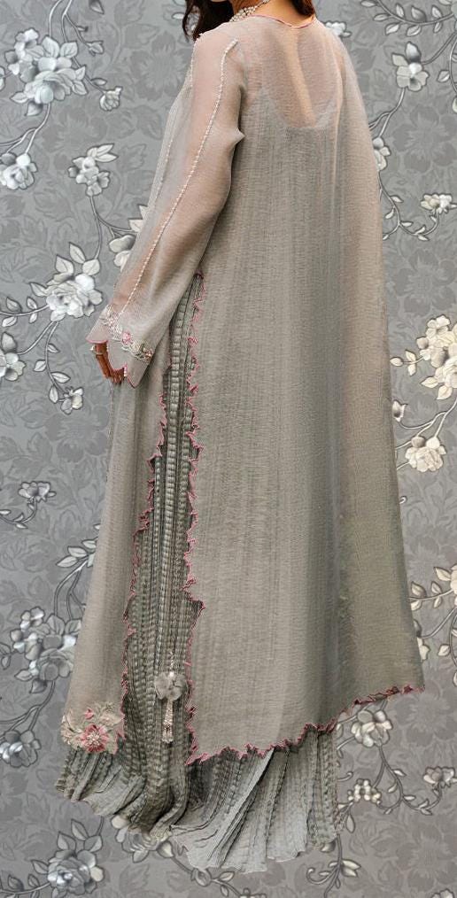 A woman wearing a long, flowing gray dress with a sheer overlay and a matching gray skirt, standing against a gray floral background.